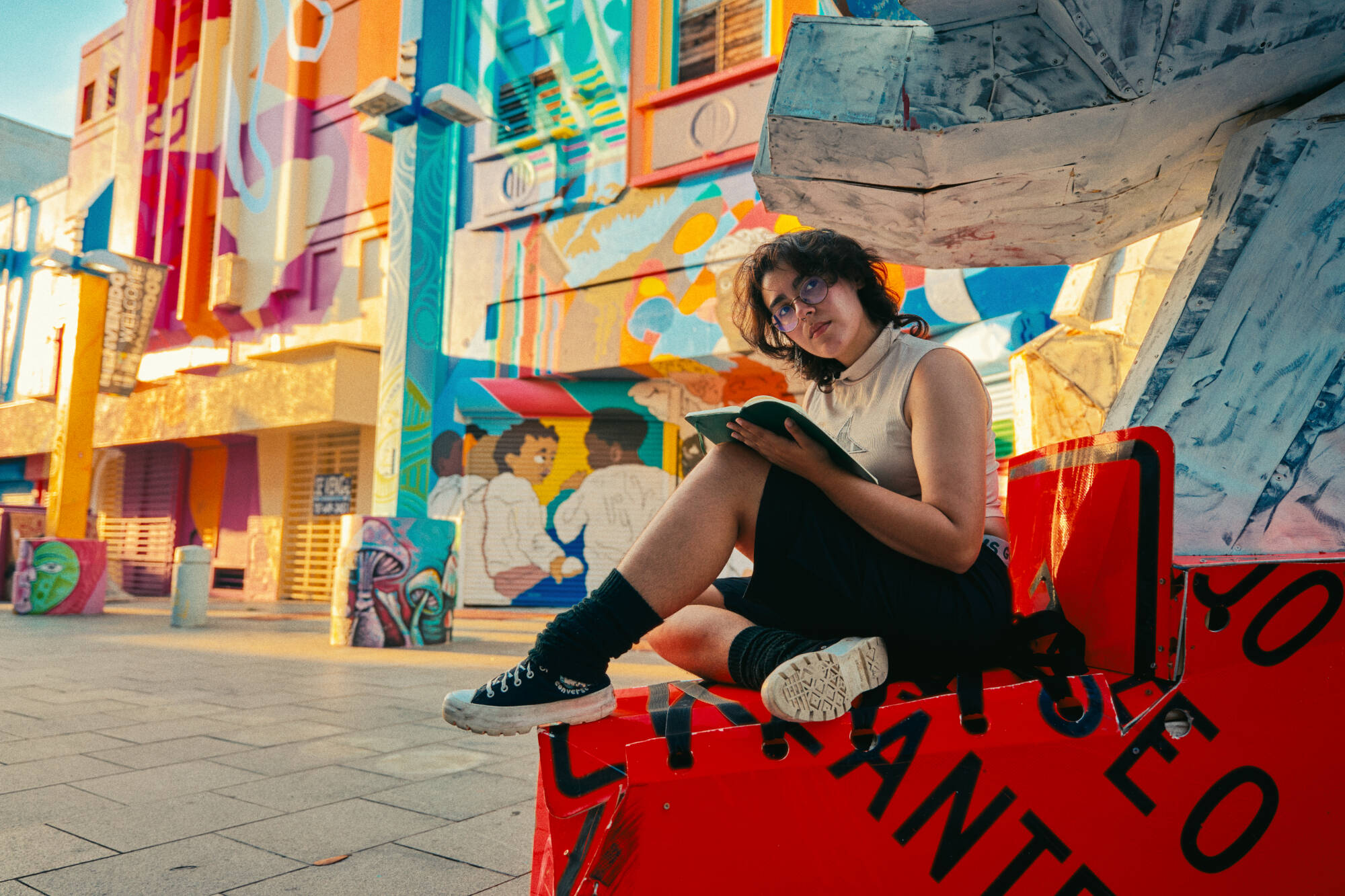 Color photograph of a person sitting, looking at a book, currounded by colorful murals.